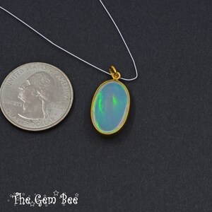 May include: Oval opal pendant with a gold-coloured bezel, suspended on a white cord. The opal displays a play of colour with green and blue hues. A US quarter is next to the pendant for size comparison. The Gem Bee logo is at the bottom.