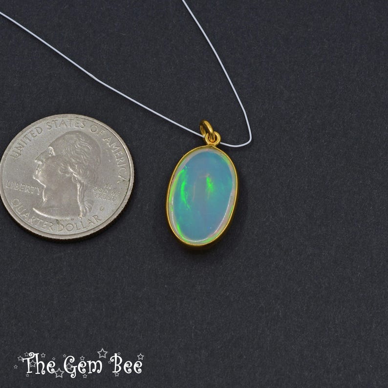 May include: Oval opal pendant with a gold-coloured bezel, suspended on a white cord. The opal displays a play of colour with green and blue hues. A US quarter is next to the pendant for size comparison. The Gem Bee logo is at the bottom.