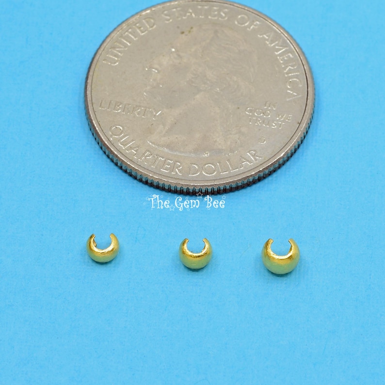 May include: Three gold-coloured metal crimp beads, each with a rounded, open-ended shape, are shown on a blue surface. A US quarter is included for size comparison.