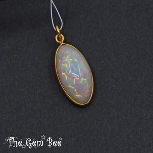 May include: Oval-shaped pendant with a gold-colored frame, featuring a white opal with iridescent flashes of color. The pendant hangs from a thin white cord. The Gem Bee logo is at the bottom.