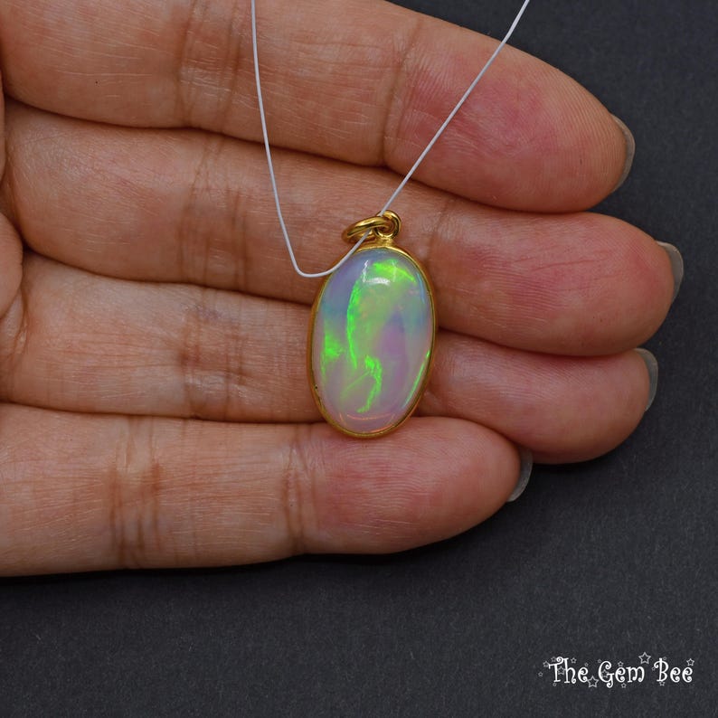May include: Oval opal pendant with a gold-coloured bezel, suspended on a white cord. The opal displays a play of colour with green and purple hues. The pendant is held against a neutral background.