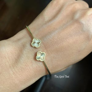 Puede incluir: Pulsera de puño abierta dorada con dos colgantes de trébol de cuatro hojas. Cada trébol tiene un centro texturizado de color crema y un contorno dorado. La pulsera se muestra en la muñeca de una persona.