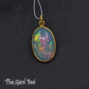 Puede incluir: Colgante de &oacute;palo ovalado con bisel y fianza dorados. El &oacute;palo muestra colores iridiscentes verdes, amarillos y rosas. El colgante cuelga de un fino cord&oacute;n blanco. El logotipo de The Gem Bee est&aacute; en la esquina inferior izquierda.
