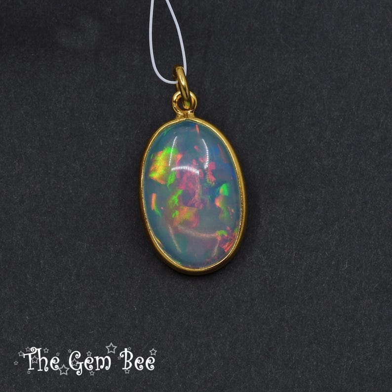 Puede incluir: Colgante de &oacute;palo ovalado con bisel y fianza dorados. El &oacute;palo muestra colores iridiscentes verdes, amarillos y rosas. El colgante cuelga de un fino cord&oacute;n blanco. El logotipo de The Gem Bee est&aacute; en la esquina inferior izquierda.