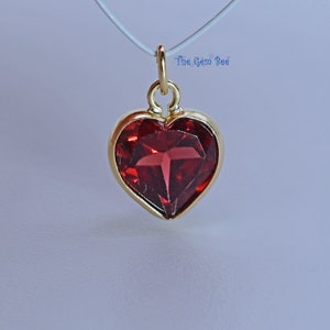 May include: A gold heart-shaped pendant with a red gemstone. The pendant is on a white string.