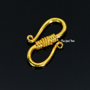 May include: A gold-colored S-hook jewelry clasp featuring a textured, coiled center section and two curved ends with small loops. The metal surface has a hammered finish. The text The Gem Bee is printed in white near the center of the clasp.