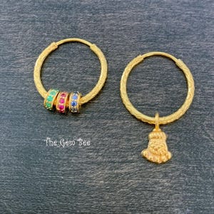 Puede incluir: Dos pendientes de aro dorados con superficies texturizadas. Un aro presenta tres peque&ntilde;as cuentas con piedras verdes, rosas y azules. El otro aro tiene un dije dorado colgante con forma de pie. El texto The Gem Bee est&aacute; impreso en blanco.