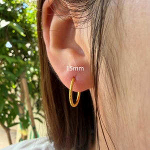 Puede incluir: Un pendiente de aro dorado con una superficie texturizada, con un di&aacute;metro de 15 mm. El pendiente es de forma circular y tiene un dise&ntilde;o de banda fina y delicada. El metal tiene un acabado mate y ligeramente granulado en la superficie.