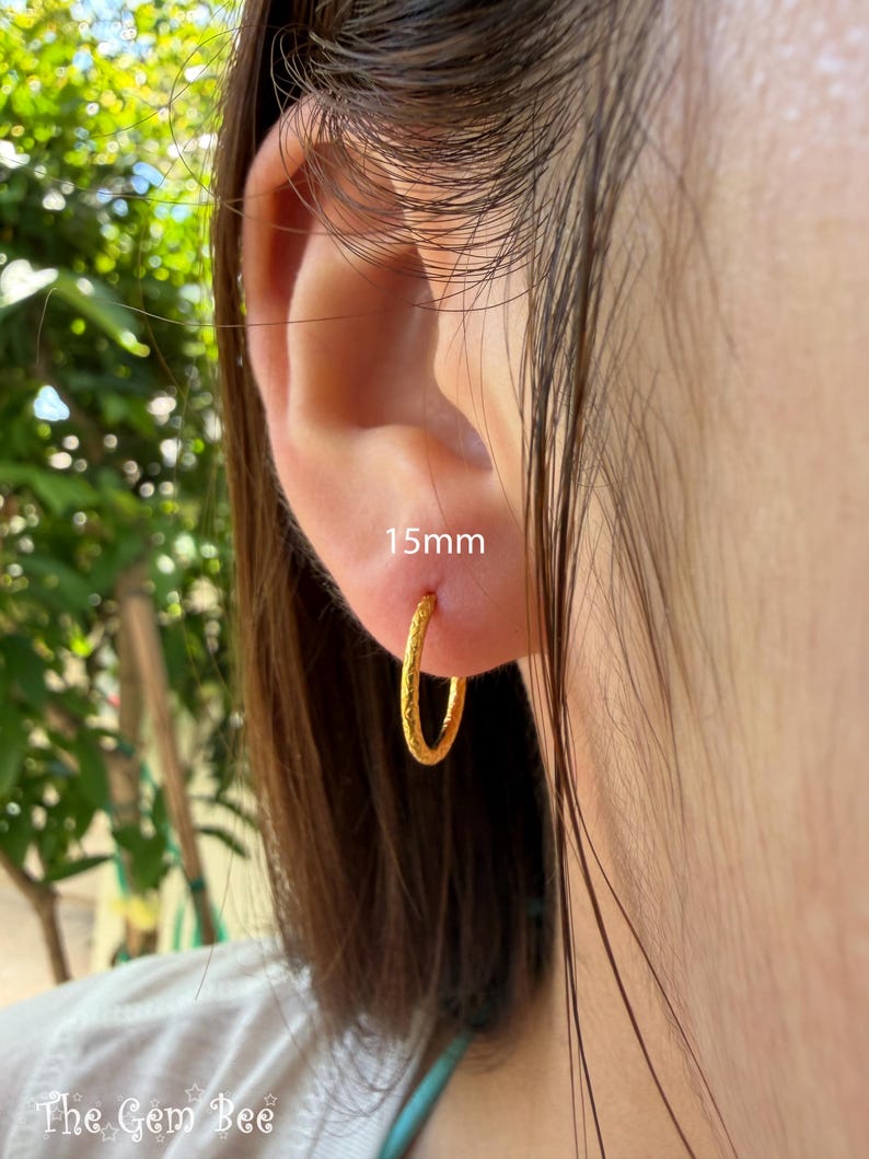 Puede incluir: Un pendiente de aro dorado con una superficie texturizada, con un di&aacute;metro de 15 mm. El pendiente es de forma circular y tiene un dise&ntilde;o de banda fina y delicada. El metal tiene un acabado mate y ligeramente granulado en la superficie.
