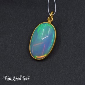 May include: Oval opal pendant with a gold-coloured bezel and bail. The opal displays iridescent colours of green, blue, and orange. The pendant hangs from a thin white cord. The Gem Bee logo is visible at the bottom.