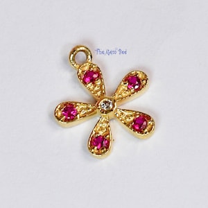 May include: Gold flower charm with five petals and a small diamond in the center. Each petal has a pink ruby gemstone.