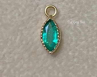 May include: A marquise-cut emerald green gemstone pendant set in a gold-toned metal bezel with a small circular jump ring at the top. The text The Gem Bee is printed in small white letters to the right of the pendant, which has a scalloped edge.