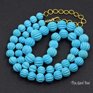 May include: A turquoise beaded necklace with a gold-coloured chain and clasp. The beads are round with a ribbed texture. The necklace is displayed on a black surface.