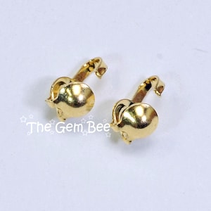 May include: Two gold-toned metal earring findings with a smooth, rounded dome shape and a curved clip mechanism. The text "The Gem Bee" is printed in white with small star outlines on the left side of the image, near the first earring finding.