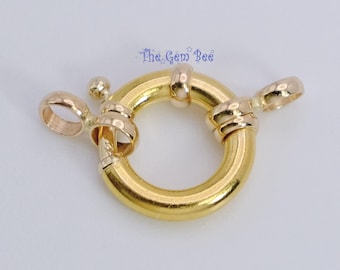 May include: A circular spring ring clasp made of polished yellow gold with a small lever mechanism and two attached jump rings. The text The Gem Bee is printed in blue above the jewelry component, which features a smooth, reflective surface.