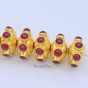 May include: A row of five gold-toned beads, each featuring a spherical shape with four embedded deep red cabochon stones arranged in a cross pattern. The text "The Gem Bee" is printed in blue below the center beads on the white background.