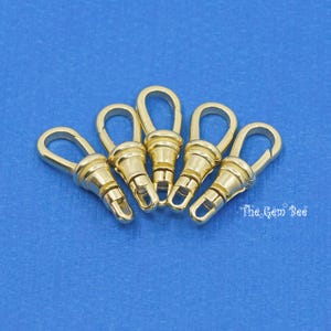 May include: Five gold-toned metal swivel clasps, also known as trigger snaps, are arranged in a semi-circle on a blue background. The clasps are a common hardware finding used in jewelry making and other crafts.