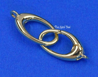 May include: A gold-toned metal jewelry clasp featuring two interlocking oval-shaped links. The design includes a small loop at each end for attachment and a spring-loaded mechanism on the larger link. The text The Gem Bee is visible.