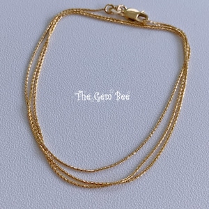 May include: A thin, gold-colored chain necklace featuring a delicate, textured link design and a small lobster claw clasp. The text "The Gem Bee" is printed in white in the center of the circular chain arrangement against a plain background.