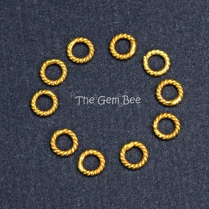 May include: A collection of ten small, circular gold-toned jump rings arranged in a circle. Each ring features a twisted rope-like texture along its surface. The text The Gem Bee is printed in white in the center of the circular arrangement.