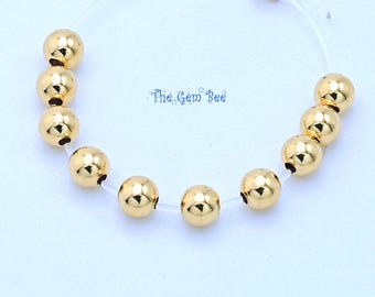 Op de afbeelding: Een cirkelvormige armband met negen gepolijste goudkleurige bolvormige kralen geregen aan een transparant, flexibel koord met een kleine goudkleurige metalen knijpkraal. De tekst The Gem Bee is in het blauw in het midden afgedrukt.