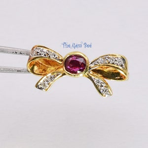 Könnte beinhalten: Ein goldener Bogenanhänger mit einem Rubin-Edelstein in der Mitte und kleinen Diamanten an den Rändern. The Gem Bee ist auf dem Bild gedruckt.