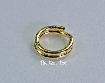 May include: A gold-toned metal split ring with a double-loop design, featuring a smooth, polished finish. The text The Gem Bee is printed in a decorative font below the ring, which is a circular jewelry component used for connecting items.