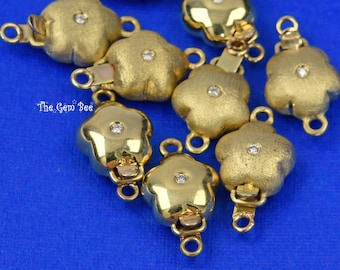 Puede incluir: Una colección de conectores para joyería en forma de flor dorada, cada uno con una pequeña piedra clara en el centro. Las piezas presentan una mezcla de acabados pulidos y mate, con pequeñas anillas circulares. El texto The Gem Bee es visible.