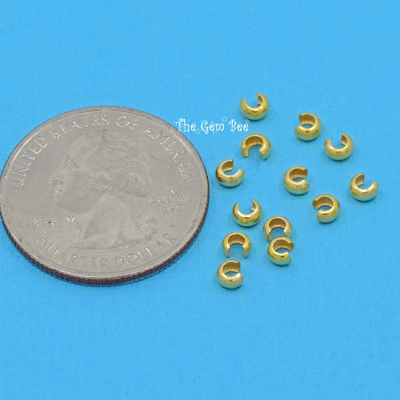 May include: A close-up of a pile of small, gold-coloured metal crimp beads. The beads are about the size of a UK 20 pence coin.