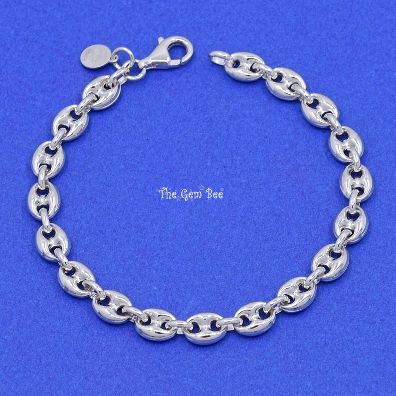 May include: A silver-colored bracelet with a lobster clasp closure. The bracelet features a series of oval-shaped links, connected to each other. A small, round charm hangs near the clasp. The Gem Bee text is visible in the image.