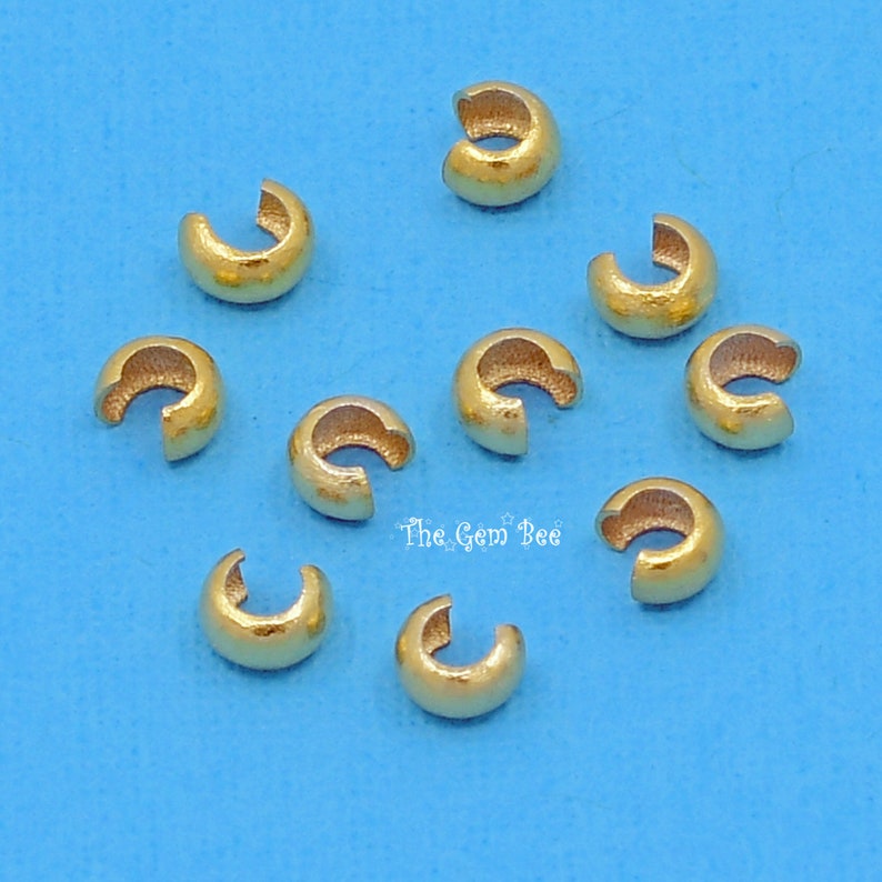 May include: Ten gold-coloured metal crimp beads, also known as crimp covers, for jewellery making. The beads are round and have a small opening on one side.