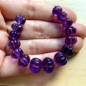 May include: A strand of deep purple, translucent amethyst beads with a carved, ribbed pumpkin-like texture. The beads are strung on a thin, clear filament. The text "The Gem Bee" is visible in the center of the arrangement of these round stones.