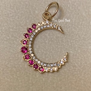 Pode incluir: Um amuleto de ouro em forma de lua crescente com uma fileira de pedras preciosas cor-de-rosa ao redor da borda. O amuleto tem um laço na parte superior para prender a um colar ou pulseira.