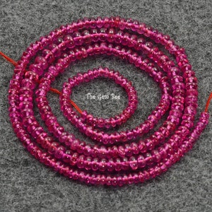 Pode incluir: Fios de contas de pedras preciosas rosa vibrante, provavelmente rubis, estão enrolados sobre uma superfície cinza. As contas variam em tamanho, com um fio menor aninhado dentro dos maiores. As contas são enfiadas em um fio vermelho.