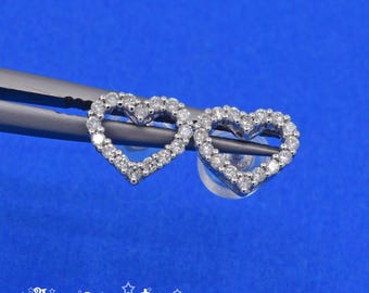 May include: A pair of silver-toned heart-shaped stud earrings, each featuring a border of small, clear, sparkling stones. The earrings are held by dark metal tweezers and include clear plastic earring backs. The text The Gem Bee is visible.