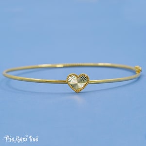 Puede incluir: Una pulsera dorada con un dije en forma de corazón. El dije de corazón tiene un diseño texturizado de rayos de sol y un borde con cuentas. La pulsera está sobre un fondo azul claro. El texto The Gem Bee está en la esquina inferior izquierda.