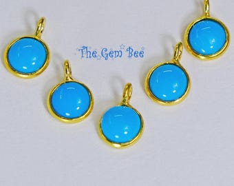 May include: Five circular charms featuring smooth, bright blue cabochons set in polished gold-tone metal frames with small top loops. The text "The Gem Bee" is printed in blue in the center of the arrangement, surrounded by small star icons.