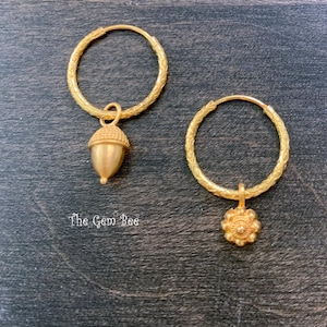 Puede incluir: Un par de pendientes de aro dorados con bandas texturizadas. Un pendiente presenta un dije colgante de bellota, mientras que el otro presenta un dije colgante con forma de flor. El texto The Gem Bee est&aacute; impreso en blanco cerca de la bellota.