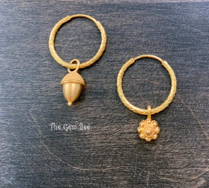 Puede incluir: Un par de pendientes de aro dorados con bandas texturizadas. Un pendiente presenta un dije colgante de bellota, mientras que el otro presenta un dije colgante con forma de flor. El texto The Gem Bee est&aacute; impreso en blanco cerca de la bellota.