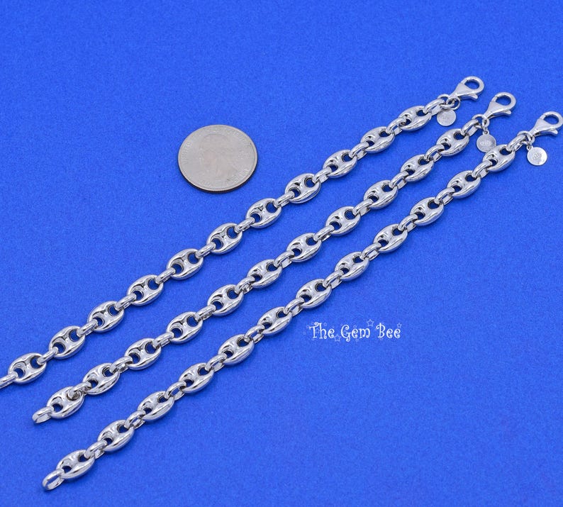 May include: Three silver-colored chain bracelets with a coffee bean link design are displayed on a blue surface. Each bracelet has a lobster clasp closure. A coin is placed near the bracelets.