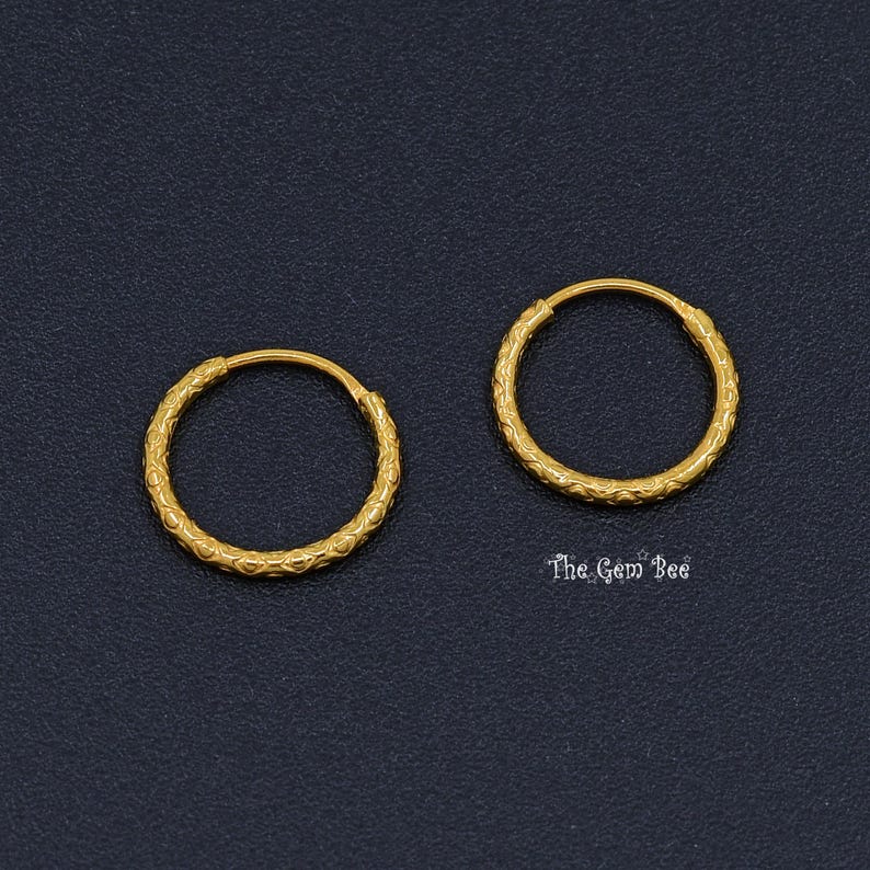 Puede incluir: Un par de pendientes de aro circulares de oro con un patr&oacute;n repetitivo y texturizado en la superficie exterior. Los aros son finos y redondos, con un acabado amarillo met&aacute;lico. El texto The Gem Bee est&aacute; impreso en blanco junto al aro derecho.