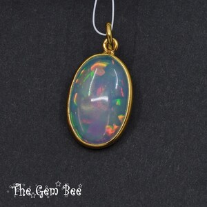 Puede incluir: Colgante de &oacute;palo ovalado con bisel y fianza dorados. El &oacute;palo muestra un juego de colores con destellos azules, verdes y naranjas. El colgante cuelga de un fino cord&oacute;n blanco sobre un fondo oscuro. El logotipo de The Gem Bee est&aacute; en la esquina inferior izquierda.