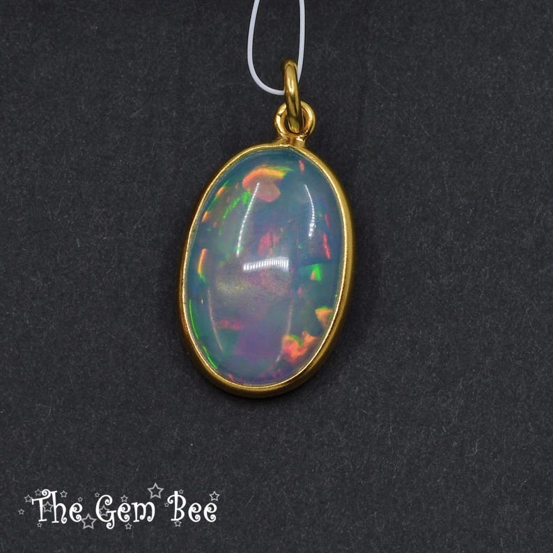 Puede incluir: Colgante de &oacute;palo ovalado con bisel y fianza dorados. El &oacute;palo muestra un juego de colores con destellos azules, verdes y naranjas. El colgante cuelga de un fino cord&oacute;n blanco sobre un fondo oscuro. El logotipo de The Gem Bee est&aacute; en la esquina inferior izquierda.