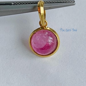 May include: A gold pendant with a round, pink gemstone. The gemstone is set in a gold bezel and has a polished, smooth surface. The pendant is suspended from a gold bail. The Gem Bee is written on the image.