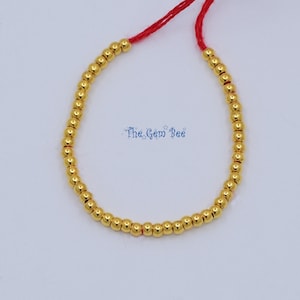 Puede incluir: Una pulsera de cuentas doradas con cierre de cordón rojo. La pulsera presenta pequeñas cuentas doradas redondas ensartadas. El texto "The Gem Bee" es visible en la imagen.