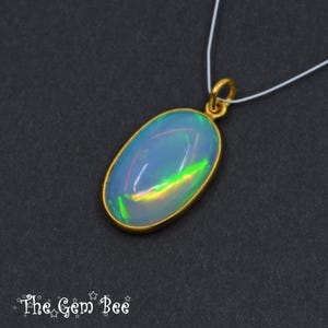 May include: Oval opal pendant with a gold-coloured bezel and bail, suspended on a white cord. The opal displays a blue and iridescent colour play with flashes of green and yellow. The Gem Bee logo is in the lower left corner.