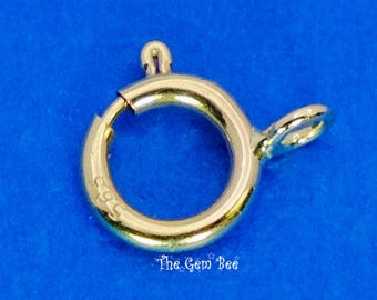 May include: A gold-toned circular spring ring clasp featuring a small attached loop on the side. The metal surface displays a stamped 585 hallmark indicating its composition. The jewelry component has a smooth, polished finish and a functional spring mechanism.