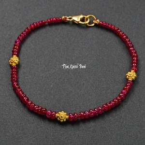 Könnte beinhalten: Ein zartes Armband aus kleinen, runden, tiefroten Edelsteinperlen. Das Design wird durch drei kleine, strukturierte, goldfarbene florale Zwischenelemente akzentuiert und mit einem goldfarbenen Karabinerverschluss geschlossen. Der Text The Gem Bee ist sichtbar.