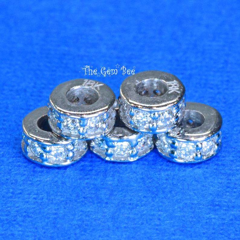 May include: Five silver-toned spacers with a row of clear cubic zirconia stones around the edge. The spacers are stacked on top of each other. The top spacer has the inscription "18K" on the side.