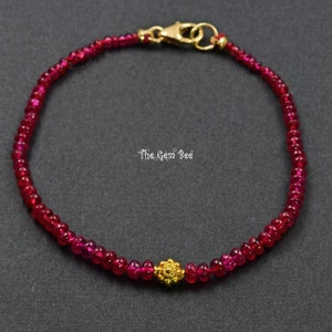 May include: A delicate bracelet featuring small, round, deep red gemstones strung together. A gold-colored, ornate bead is centered on the bracelet. The clasp and connecting hardware are also gold-colored. The bracelet is displayed on a dark gray surface.